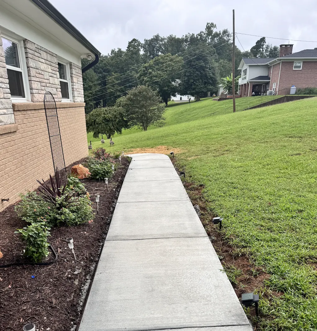 Straight concrete walkway with evenly spaced control joints beside a stone-front home, bordered by a planted bed and path lights &mdash; The Foothills Concrete Company