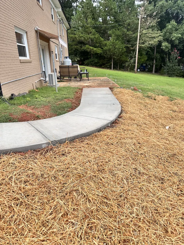 Curved concrete walkway with tooled control joints running alongside a brick home to a rear patio, edged with fresh pine straw &mdash; The Foothills Concrete Company