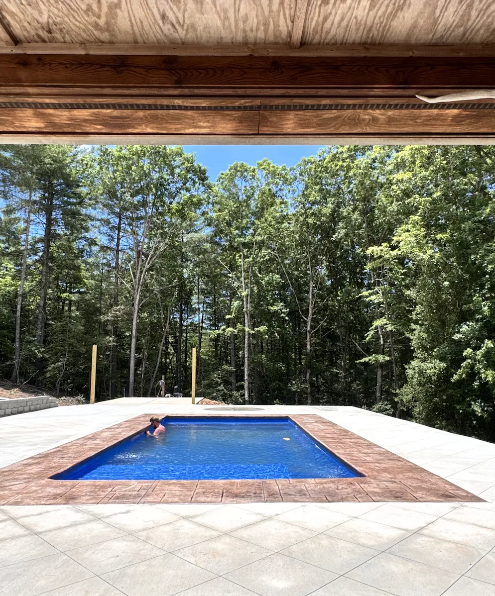 View from a covered porch across a wide stamped concrete pool deck to a rectangular pool with rust-toned stamped coping, a swimmer in the water and mature woods beyond &mdash; The Foothills Concrete Company